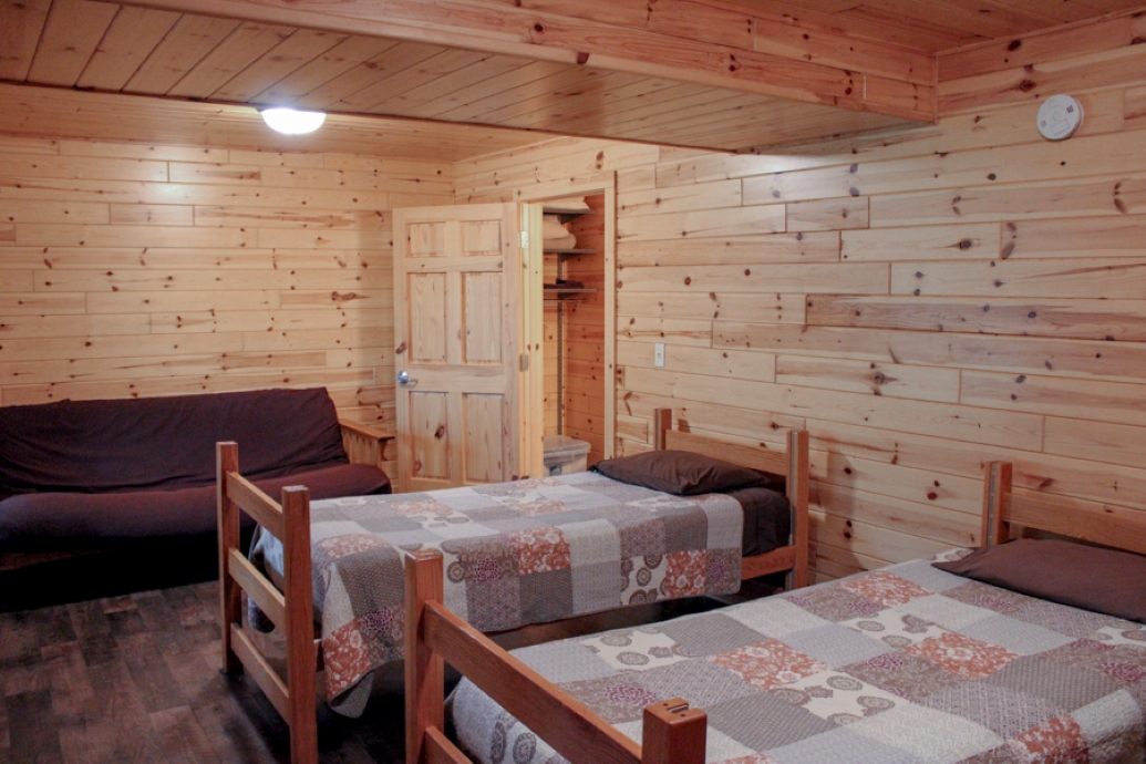 Cabin basement including two twin beds with quilts, a futon, and a door in the corner that leads to 