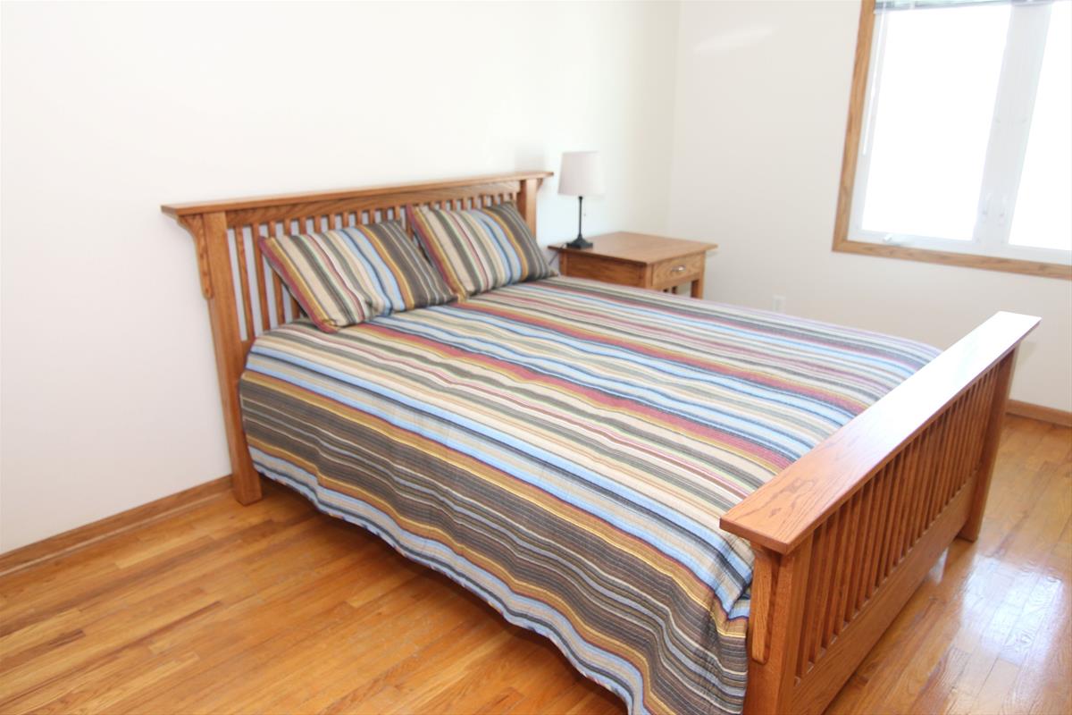 Main bedroom including queen bed and quilt, mission wood bedframe, nightstand and lamp, and large wi